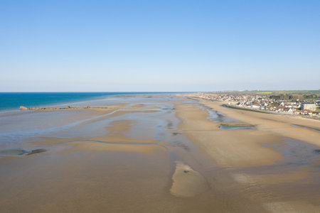 Drone view of the expansive sandy shoreline and shallow tidal pools at Gold Beach near Asnelles, Normandy, with a coastal village and clear blue sky on a bright day.の写真素材