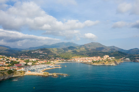 Wide aerial view of Banyuls-sur-Mer featuring a bustling harbor, terracotta-roofed buildings, and lush mountains rising in the background beneath a sky with scattered clouds.の写真素材