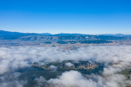Aerial panorama of the island on Lake Ioannina partially veiled by low clouds, with the city of Ioannina and the Pindus mountains rising in the distance under a vivid blue sky in Epirus, Greece. The composition highlights the interplay of atmospheric layers, natural textures, and urban landscape.の写真素材