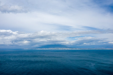 Expansive view across the deep blue waters of the Bay of Naples toward Mount Vesuvius, partially shrouded by dramatic clouds. Soft natural light highlights the textured surface of the sea and the layered sky.の写真素材