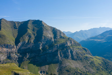 Majestic green and rocky cliffs rise sharply under a clear blue sky in the Pyrenees near Gavarnie Gedre. Layers of distant peaks fade into the horizon, creating a tranquil and expansive alpine scene.の写真素材