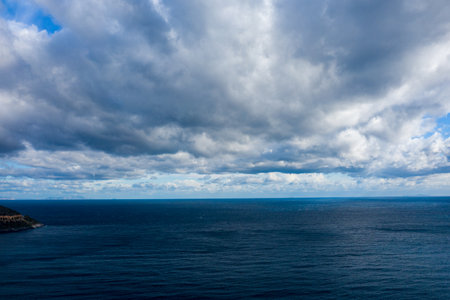 Expansive view of deep blue water stretching to the horizon under a sky filled with thick, textured clouds. The moody atmosphere is enhanced by shifting light and subtle shadows on the open sea.の写真素材