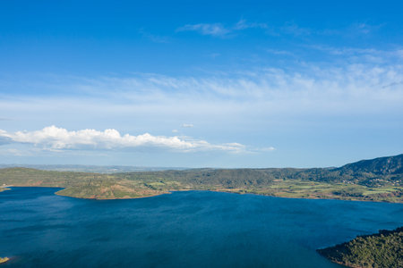 Expansive aerial view of Lac de Salagou with deep blue water, rolling green hills, and farmland stretching beneath a bright blue sky with scattered clouds.の写真素材