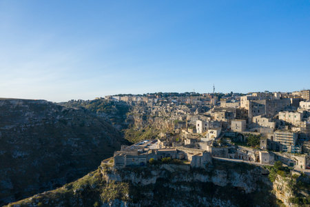 Wide view of Matera, Italy, featuring historic stone structures clustered atop a rugged plateau with steep cliffs, illuminated by clear daylight against a vivid blue sky.の写真素材