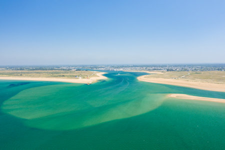 Aerial perspective of vivid turquoise tidal waters flowing between golden sandbanks at Barre dEtel, Brittany. The scene features a clear blue sky, expansive coastline, and distant town on a bright summer day.の写真素材