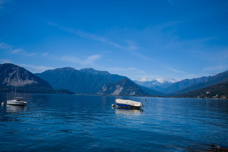 Small boats float on the calm blue waters of Lake Maggiore in northern Italy, framed by rugged alpine peaks and forested hills under a clear sky. The tranquil scene highlights the contrast between the deep water and the dramatic mountain landscape.の写真素材