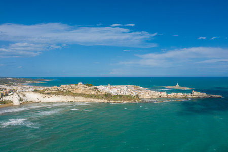 Expansive aerial panorama of Vieste, Italy, featuring white limestone cliffs, a historic old town, and a curved sandy beach along the Adriatic Sea. The scene is bathed in bright sunlight beneath a vivid blue sky with scattered clouds, highlighting the dramatic coastline and Mediterranean atmosphere.の写真素材