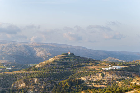 A small white chapel crowns a terraced hill surrounded by olive groves and rugged mountains in...