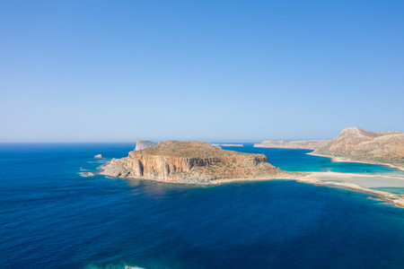 A striking rocky headland rises above deep blue waters at Balos Beach, Crete, Greece. The scene is...
