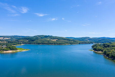 Wide view of Panneciere-Chaumard reservoir with calm blue water surrounded by rolling forested hills and open farmland under a clear summer sky in Burgundy. The image captures tranquil aquatic scenery and the gentle contours of the Morvan region.の写真素材