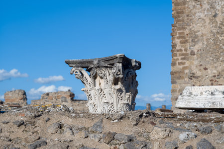Detailed stone carving of a Corinthian column capital rests atop weathered volcanic masonry in the archaeological site of Pompeii, Italy. Bright sunlight and a vivid blue sky highlight the intricate relief and ancient textures of this historic Roman city.の写真素材