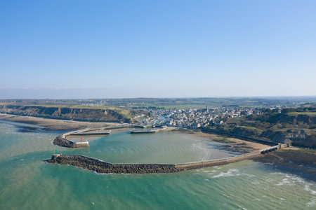 Wide drone view of Port en Bessin showing the harbor, protective sea walls, and surrounding cliffs under clear blue skies. The tranquil water and coastal village create a peaceful maritime scene.の写真素材