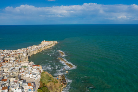 Wide aerial panorama of Vieste, Italy, showing the historic old town and cathedral perched on a rocky promontory jutting into the turquoise Adriatic Sea. The scene features whitewashed buildings, textured waves, and a vast expanse of blue sky.の写真素材