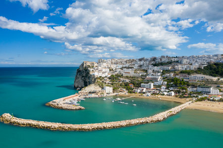 Aerial view of Peschicis marina with turquoise water, stone breakwaters, and a hillside of whitewashed buildings set against a dramatic sky with scattered clouds.の写真素材