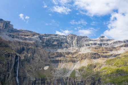 Expansive stratified cliffs dominate the landscape in Cirque de Gavarnie, with a delicate waterfall and patches of green grass beneath a partly cloudy sky.の写真素材