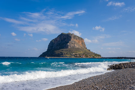 Turquoise waves break on a stony pebble beach with Monemvasia island rising steeply in the background under a clear blue sky. The rugged rock formation and vivid sea create a dramatic coastal scene.の写真素材