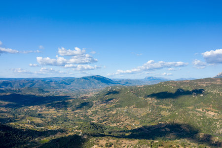 Wide aerial view of rolling hills and patchwork farmland stretching toward distant blue mountains under a clear sky with scattered clouds near Orgosolo, Sardinia.の写真素材