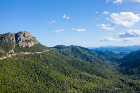 Elevated road curves along a rugged green mountain ridge in Sardinia, surrounded by dense forest and dramatic rocky cliffs under a bright blue sky with scattered clouds.の写真素材