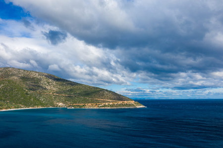 A sweeping coastal headland covered in green vegetation meets deep blue waters beneath a sky filled with thick, textured clouds. The rugged shoreline and vivid colors evoke a sense of wild Mediterranean beauty.の写真素材