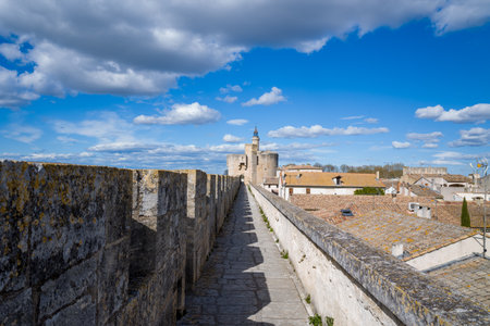 Long perspective along ancient stone ramparts overlooking rustic tiled rooftops, with the iconic medieval tower of Aigues-Mortes under a vivid blue sky and scattered clouds.の写真素材