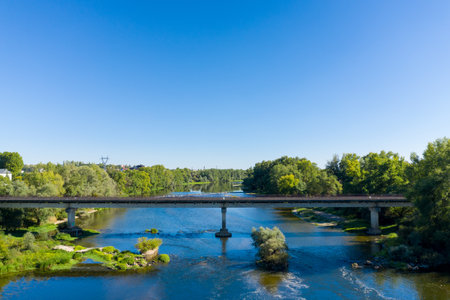 Concrete bridge spans a calm river surrounded by dense green trees and grassy banks under a clear blue sky. The tranquil scene features natural reflections, flowing water, and the peaceful rural landscape typical of the Loire Valley near Imphy in central France.の写真素材
