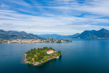 Aerial panorama of Isola Madre surrounded by deep blue waters of Lake Maggiore, with lush gardens and a historic villa in the foreground. The scene features distant alpine mountains, scattered clouds, and the lakeside towns of northern Italy.の写真素材