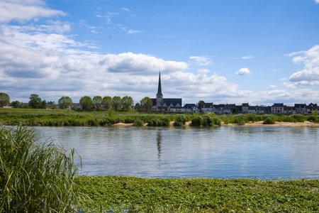 Traditional village buildings and a tall church steeple line the horizon, mirrored in the calm waters of the Loire river with lush green banks under a bright blue sky.の写真素材