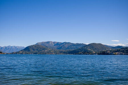 Wide view across Lake Orta in northern Italy, featuring deep blue water and gentle mountain slopes beneath a cloudless sky. The distant shoreline is dotted with small villages and lush green hills, creating a peaceful natural scene.の写真素材