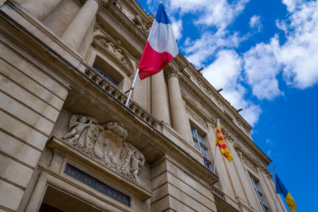 Low angle view of the ornate stone facade of the Arles city hall, featuring classical columns, sculpted reliefs, and vibrant flags against a bright blue sky with scattered clouds.の写真素材