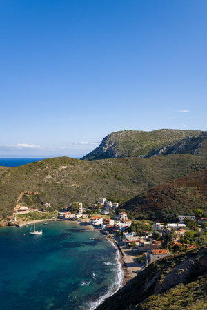 A small coastal village with whitewashed buildings lines the turquoise bay of Porto Kagio, Greece, surrounded by rugged hills under a vast clear blue sky. Sunlight highlights the textures of the water and the natural landscape, creating a peaceful Mediterranean scene.の写真素材