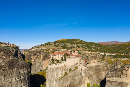Varlaam Monastery stands atop massive rock pillars in Meteora, Greece, with terracotta roofs contrasting against rugged stone and a vivid blue sky. The landscape features rolling hills, scattered trees, and dramatic autumn colors.の写真素材