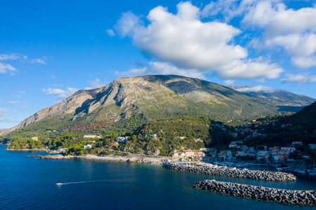 Picturesque harbor with stone breakwaters and a small boat, set against lush hills and dramatic rocky peaks under a bright blue sky with scattered clouds.の写真素材