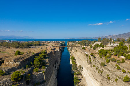 Expansive aerial view of the Corinth Canal in Greece, with steep limestone cliffs and a distant bridge opening toward the sparkling blue sea. Lush green pines and dry terrain frame the dramatic waterway beneath a clear sky.の写真素材