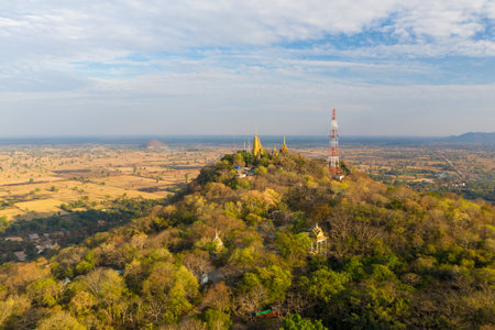Aerial panorama of golden pagodas and temple structures perched atop the forested summit of Phnom Sampov, overlooking expansive dry-season plains and rural fields near Battambang, Cambodia. The scene features intricate Khmer architecture, a communications tower, and a wide agricultural landscape beneath a sky with scattered clouds.の写真素材