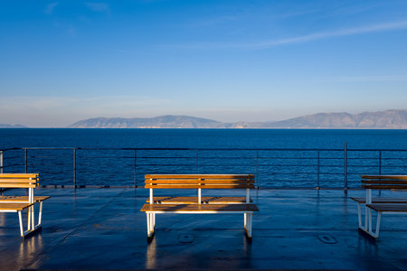 Sunlit wooden benches on a ferry deck overlook the deep blue Ionian Sea, with distant mountains rising across the horizon under clear morning skies. The scene features crisp reflections, geometric lines, and a tranquil maritime atmosphere.の写真素材