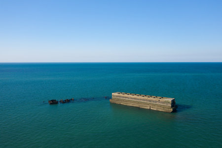 Minimalist aerial view of a massive rectangular concrete structure and scattered ruins from a historic harbor floating in tranquil turquoise water off the coast of Asnelles, Normandy, under a clear blue sky.の写真素材