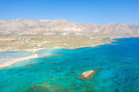 Aerial view of Elafonisi beach in Crete, Greece, featuring a vivid turquoise lagoon, rocky outcrop, and expansive arid coastline beneath distant mountains. The scene is bright, sunlit, and naturally vibrant.の写真素材