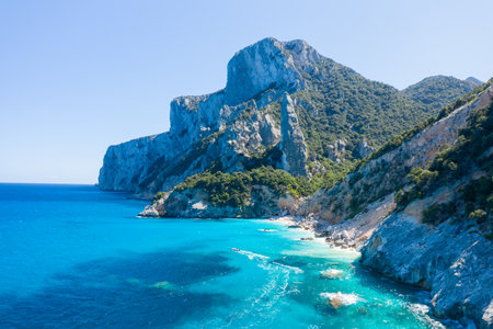 Jagged limestone cliffs tower above vibrant turquoise sea and lush green vegetation at Cala Goloritze, Baunei, Sardinia. Bright sunlight enhances the dramatic coastal landscape and crystal-clear water.の写真素材