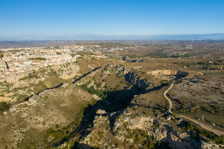 Aerial view of rugged limestone cliffs and winding dirt roads on the outskirts of Matera, Italy, with the city blending into a vast rural landscape under a clear blue sky.の写真素材