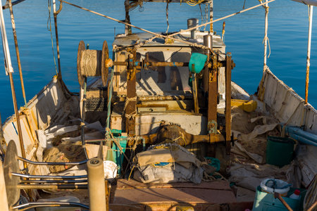 Sunlight illuminates the worn deck and tangled fishing gear inside a rustic boat moored in Kilada, Greece. The scene showcases maritime textures, aged equipment, and calm blue water in the background.の写真素材