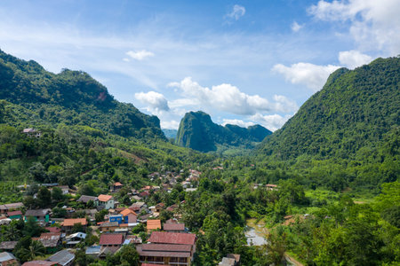A panoramic view of Nong Khiaw, Laos, reveals a vibrant village surrounded by lush green limestone mountains and dense tropical forest under a clear blue sky. The scene captures traditional rooftops, winding roads, and dramatic natural formations in northern Laos.の写真素材