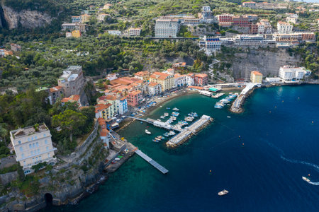 High-angle panorama of Sorrento, Italy, with vibrant houses clustered around a small marina and dramatic cliffs rising behind. The scene features turquoise Mediterranean water, lush greenery, and a lively coastal atmosphere.の写真素材