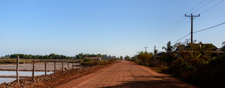 A straight unpaved road of reddish soil runs parallel to salt evaporation ponds and rustic fencing near Kampot, Cambodia. The scene features clear blue sky, scattered houses, and utility poles, capturing the open landscape and tranquil rural atmosphere.の写真素材
