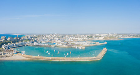 High-angle panoramic photo of Granville marina, featuring a sweeping stone breakwater enclosing turquoise water dotted with small boats. The coastal town stretches into the distance under bright sunlight and a clear blue sky.の写真素材