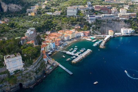 High-angle panorama of Sorrento, Italy, with vibrant houses clustered around a small marina and dramatic cliffs rising behind. The scene features turquoise Mediterranean water, lush greenery, and a lively coastal atmosphere.の写真素材
