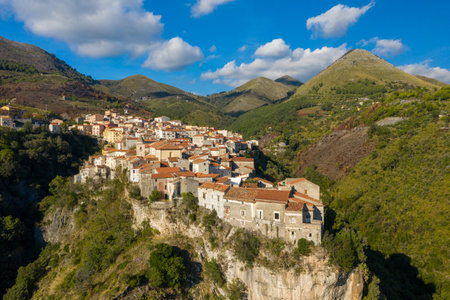 Elevated view of Tortora, Italy, showing sunlit stone houses with terracotta roofs clustered along a dramatic cliff edge, surrounded by lush green hills and rugged mountains beneath a blue sky with scattered clouds.の写真素材