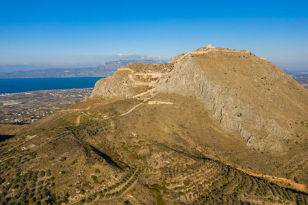 Drone view of the imposing Acrocorinth citadel standing on a steep, rocky hill above olive groves and farmland, with the deep blue gulf and distant mountains in Greece under a clear sky.の写真素材
