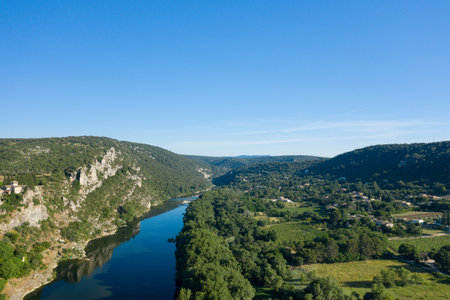 Drone shot of a winding river flowing through a lush green valley with rocky hillsides and scattered rural houses near Aigueze under a clear blue sky.の写真素材