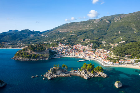 Wide aerial view of Parga in Epirus, Greece, displaying a vibrant coastal village with red-roofed houses, a historic hilltop fortress, and rocky islets surrounded by turquoise waters. The Mediterranean landscape is framed by densely forested hills and rugged mountains under a clear blue sky.の写真素材