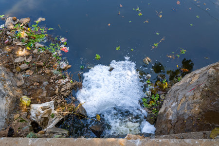 Foamy wastewater flows from a concrete drain into a river in Alappuzha, India, surrounded by rocks, litter, and floating debris. The scene highlights pollution and environmental impact under natural daylight.の写真素材